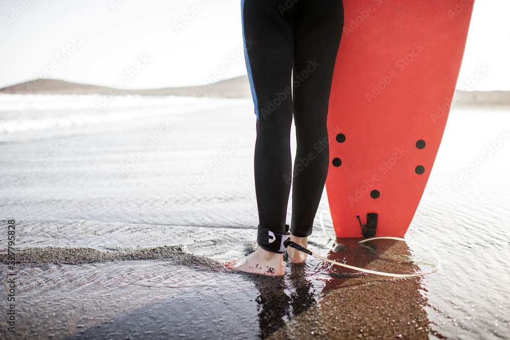 Surfer standing with surfboard on the sandy beach on a sunset, cropped view focused on womans legs