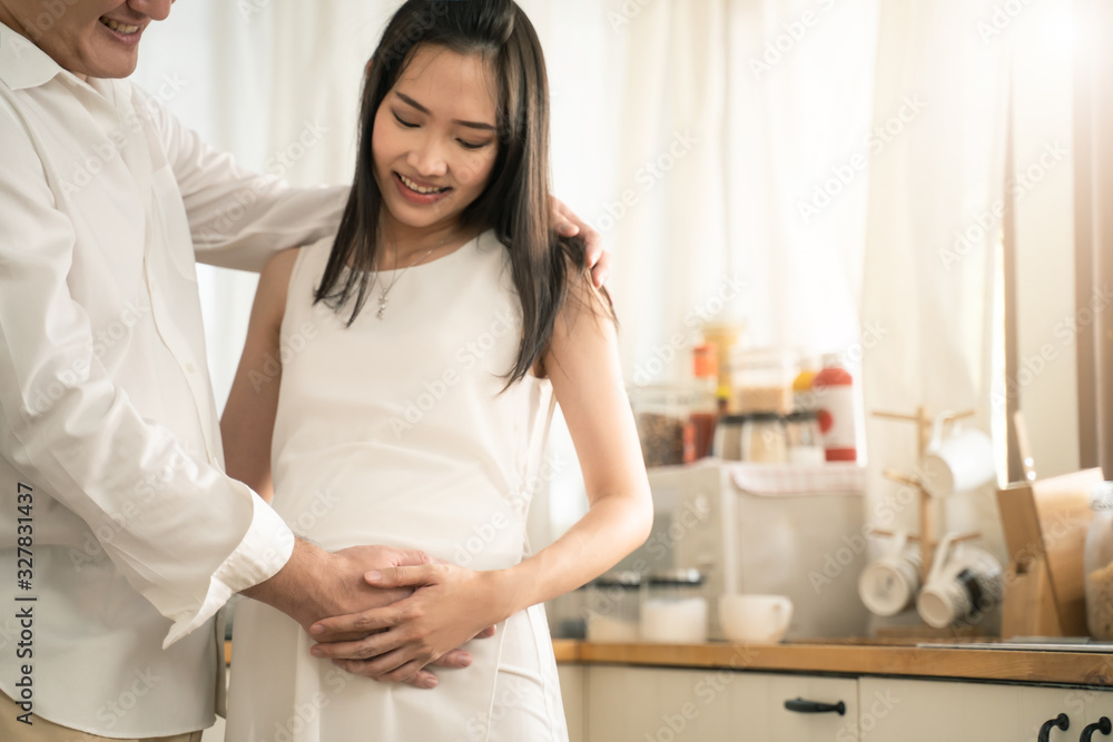 Asian man and pregnancy woman stand in kitchen at home. Father holding mother tummy feeling love exp