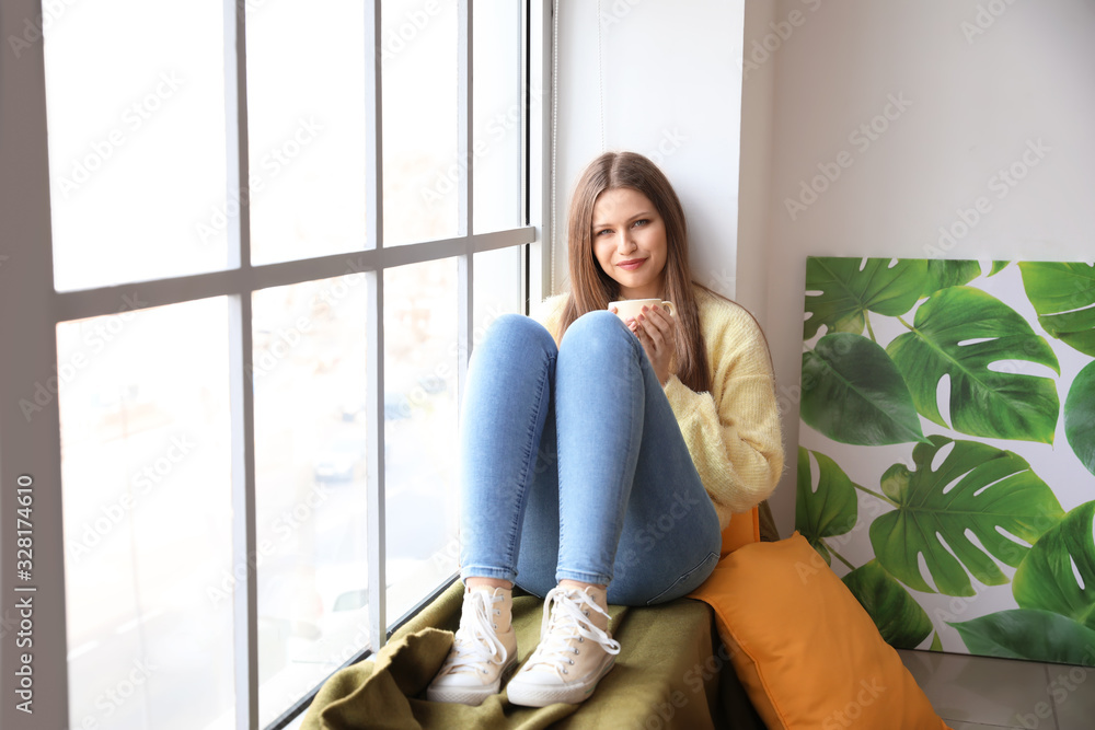 Beautiful young woman with cup of tea sitting on window sill at home