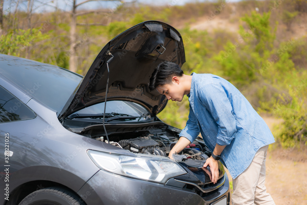 Asian young unhappy man inspecting broken car engine  in front of the open hood  broken down car On 