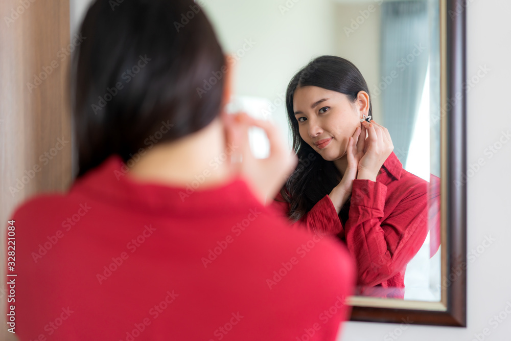 Beautiful Asian Woman wearing red dressed putting star earring looking in mirror in her bedroom at h