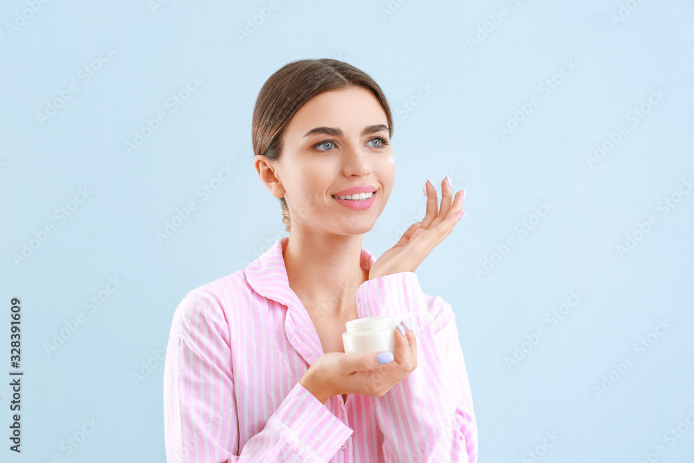 Beautiful young woman applying cream on her face against color background