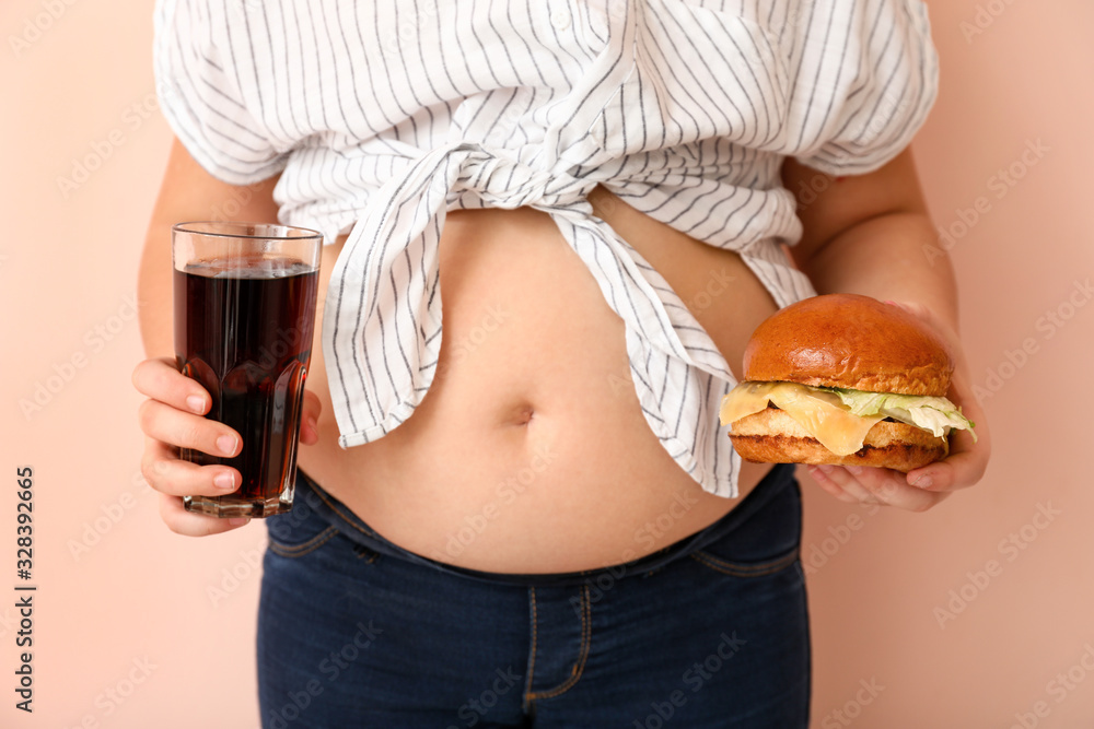 Overweight girl with unhealthy burger and drink on color background