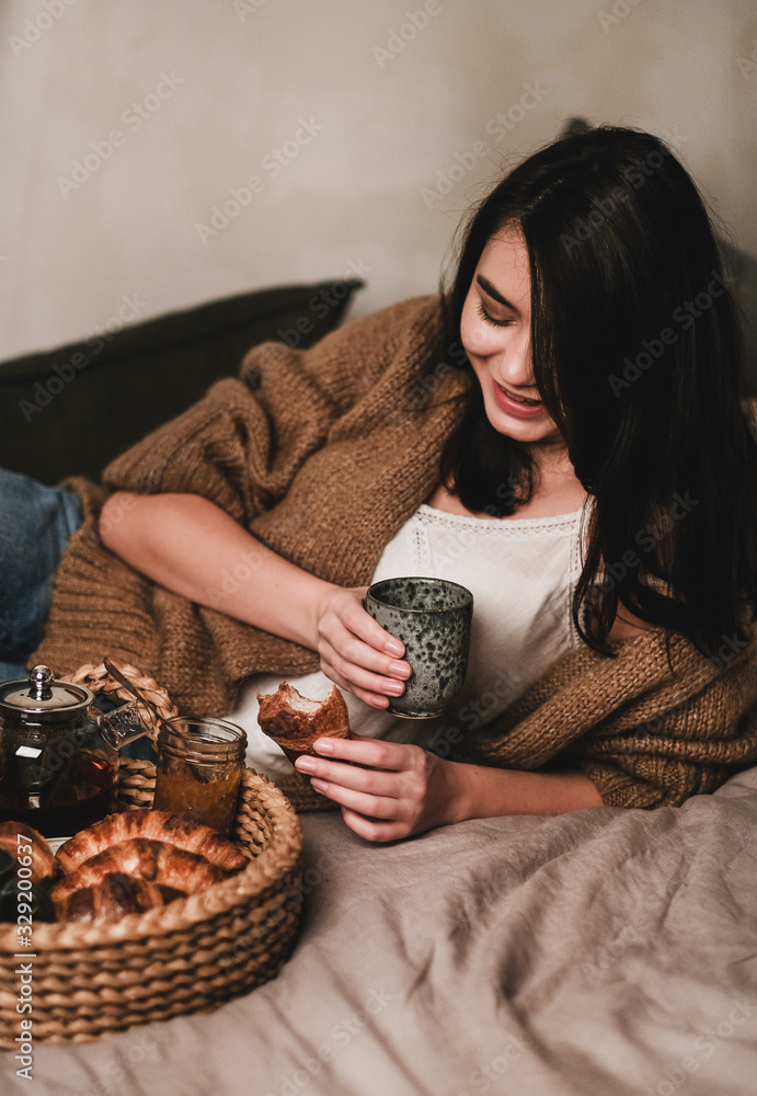 穿着舒适家居服的年轻微笑的深色头发女人在床上用茶和玉米饼吃早餐