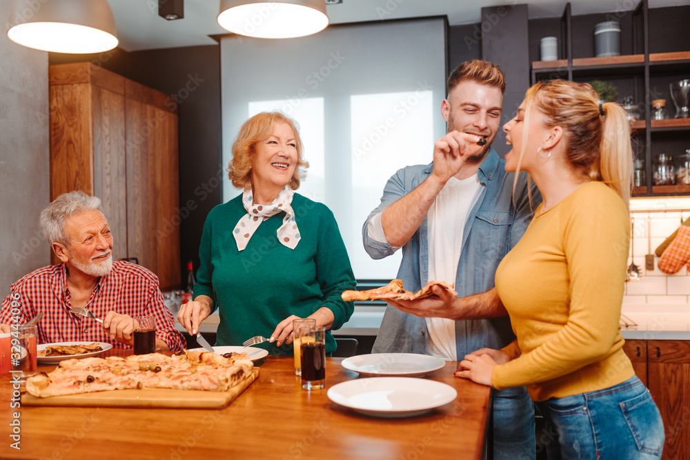 Family is eating pizza in the kitchen. Young man is giving an black olive to his girlfriend.