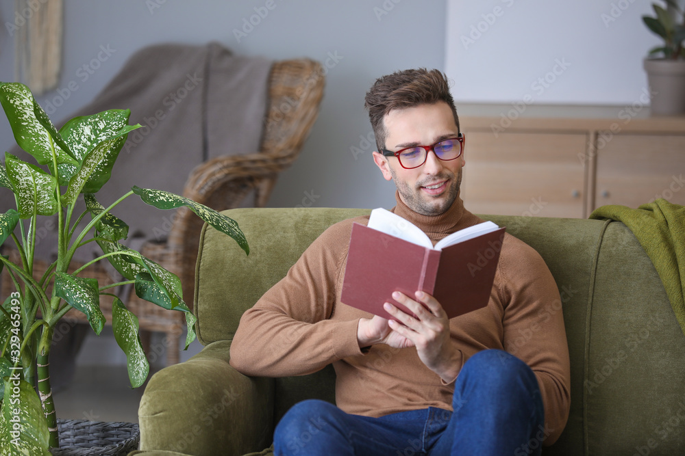 Young man with stylish eyeglasses reading book at home