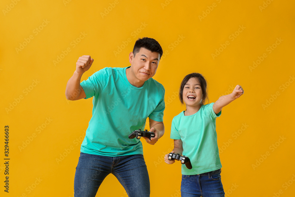 Asian man and his little daughter playing video game on color background