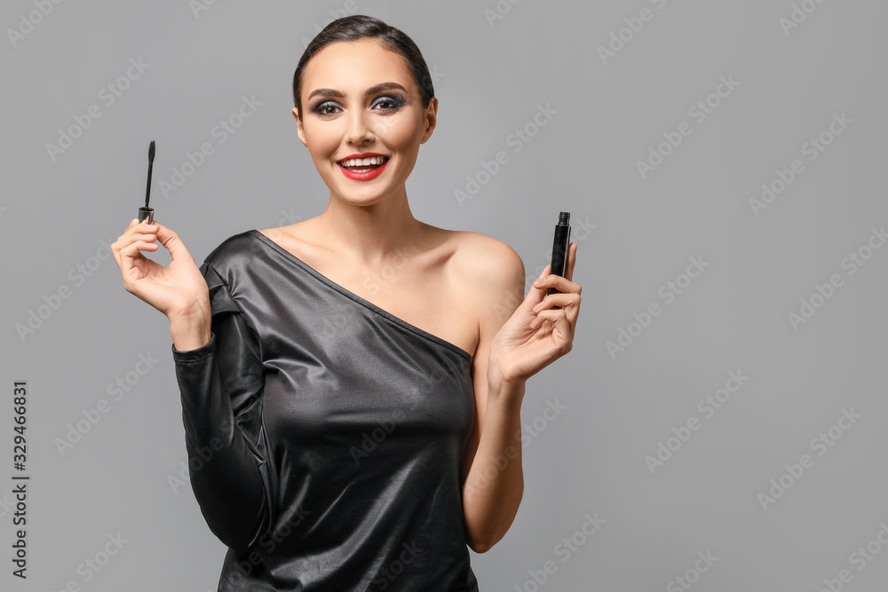 Beautiful young woman applying makeup against grey background