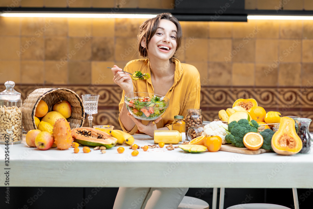 Portrait of a young cheerful woman eating salad at the table full of healthy raw vegetables and frui