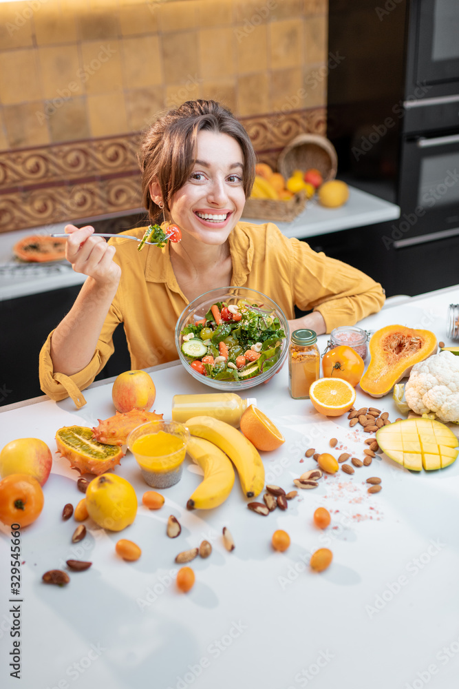 Portrait of a young cheerful woman eating salad at the table full of healthy raw vegetables and frui