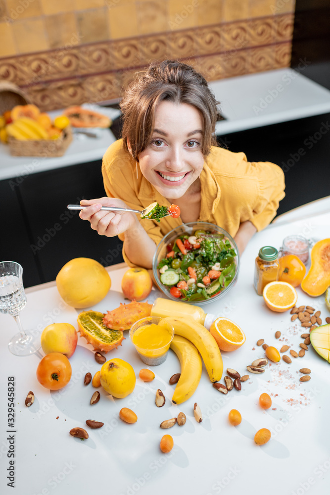 Portrait of a young cheerful woman eating salad at the table full of healthy raw vegetables and frui