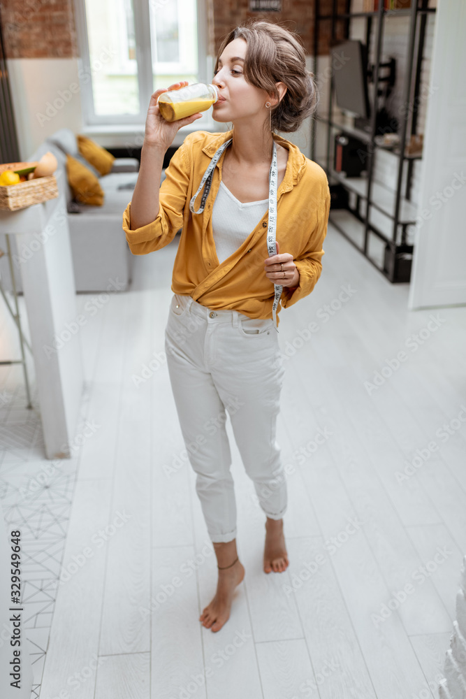 Full body portrait of a young and cheerful woman with juice and measuring tape at home. Weight loss 