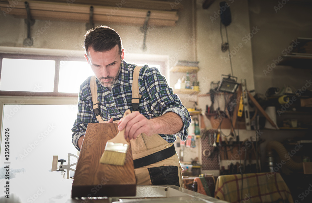 Craftsman applies varnish on wooden board by paintbrush in his carpentry workshop