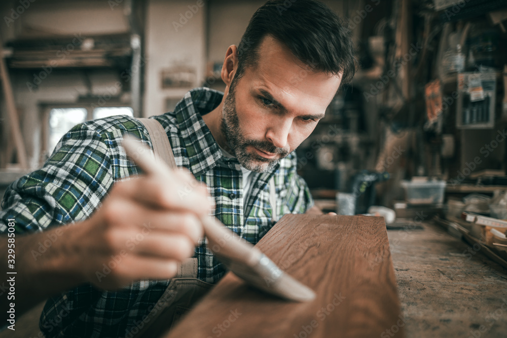 Craftsman applies varnish on wooden board by paintbrush in his carpentry workshop