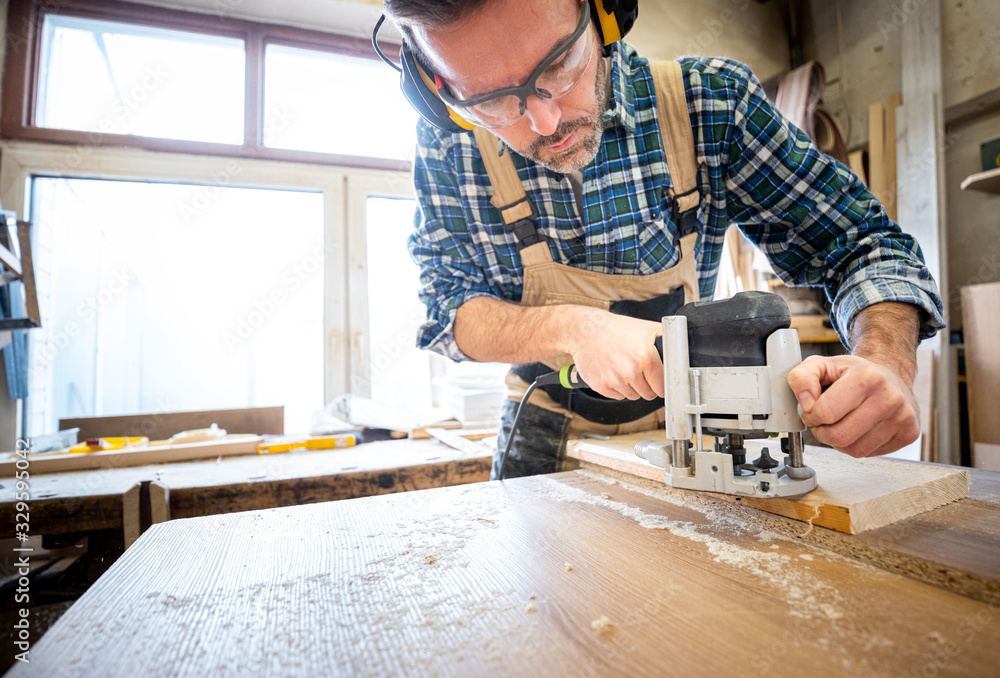 Carpenter holds milling machine and and working at wooden board in woodworking workshop