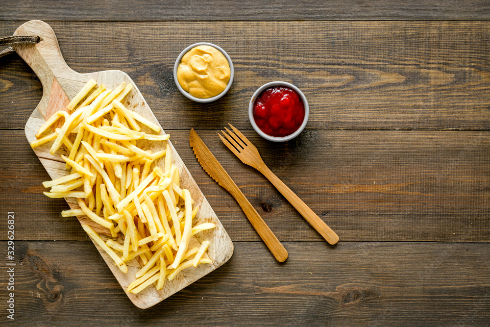 Fast food restaurant concept. French fries on cutting board near sauces on wooden table top-down cop