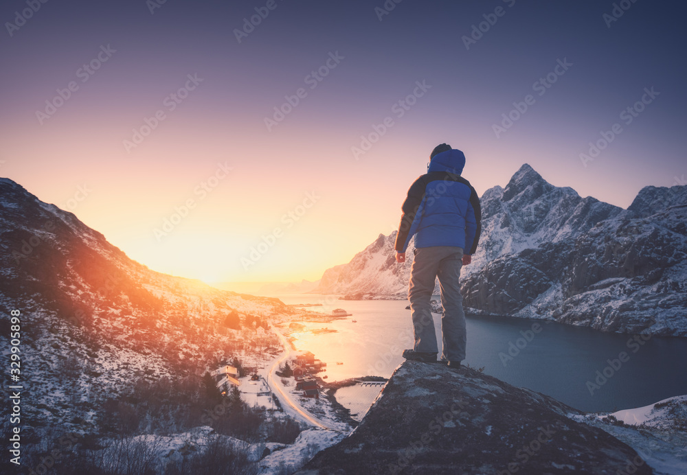 Man is standing on the mountain peak against snowy mountains, fjord at colorful sunset in winter. Yo