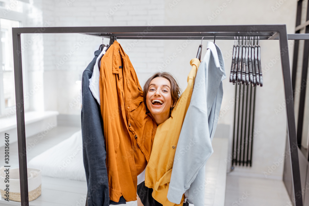 Portrait of a young and cheerful woman choosing casual clothes to wear, standing between hangers and