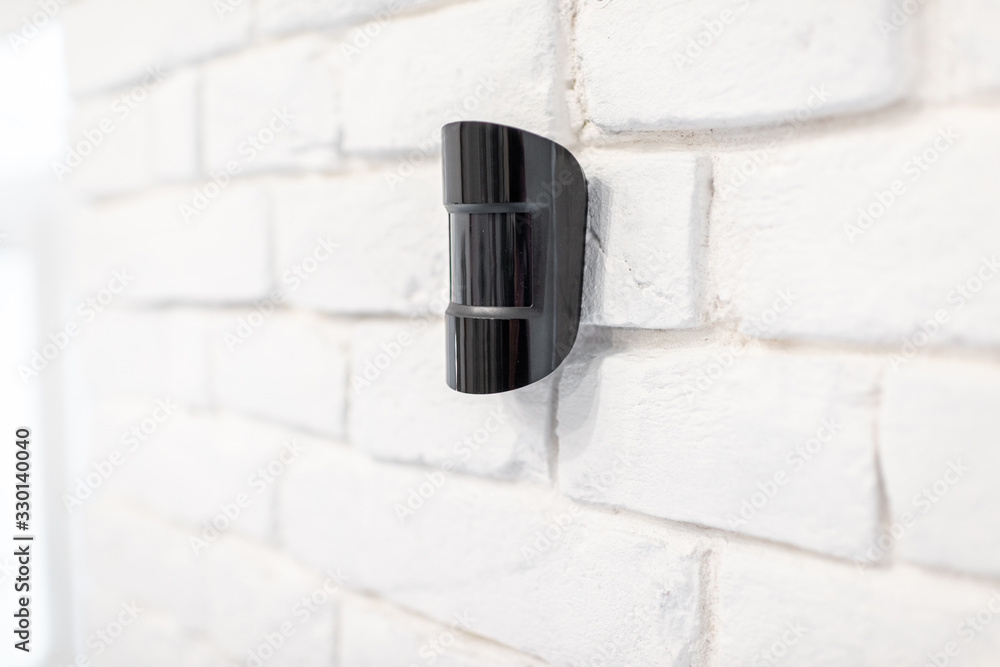 Black wireless motion sensor of alarm system mounted on the white brick wall indoors