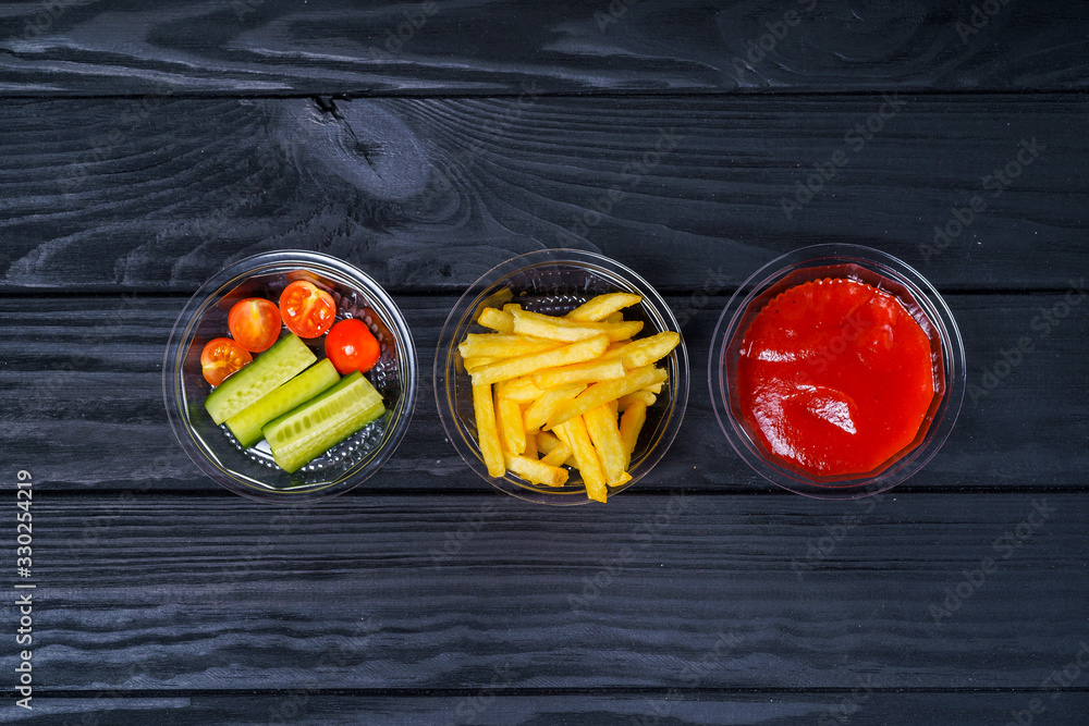 Containers with fast food on dark wooden table. French fries, ketchup and vegetables. Delivery servi