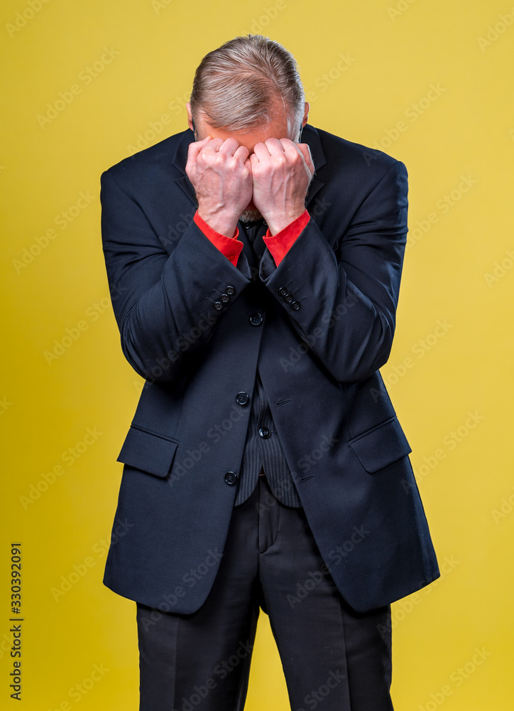 Portrait of a sad man standing on yellow background. Senior man covers face with hands. Depression o