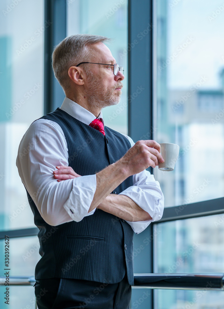 Senior businessman wears white shirt. Drinking coffee near big window with city view . Photo from th