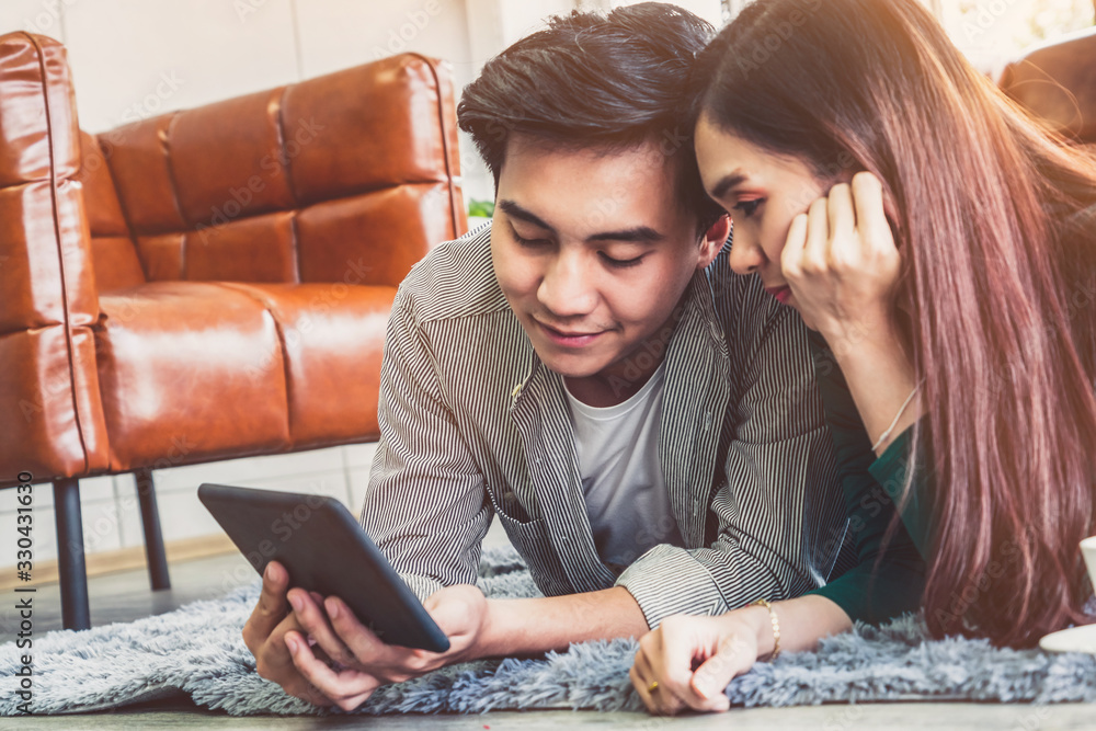 Happy Asian couple use tablet while lying down on carpet at living room floor. Love and relationship