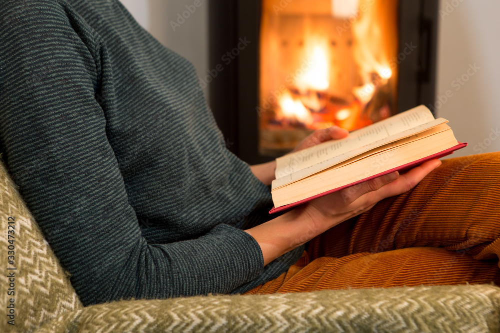 Young woman is sitting on a chair near a fireplace and reading a book, holding a cup of tea