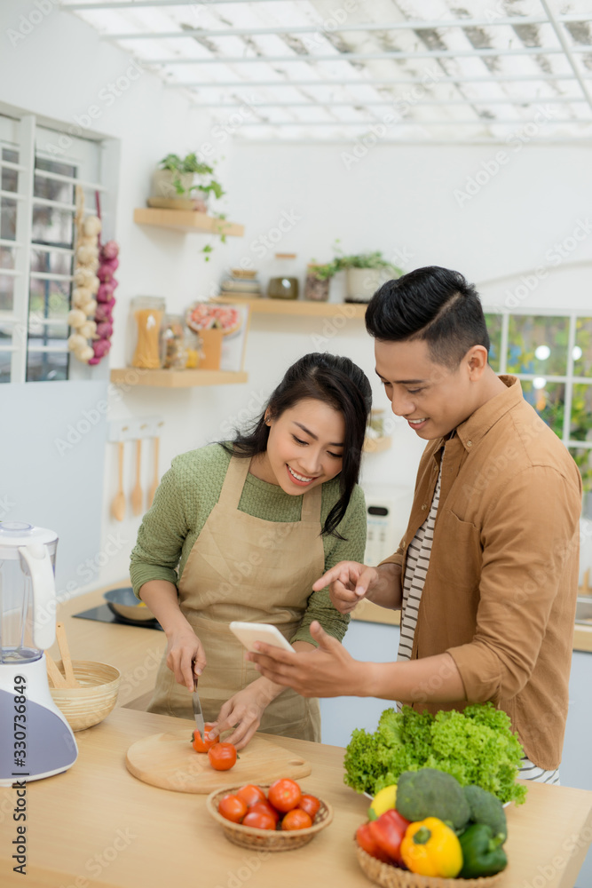 一对漂亮的年轻夫妇在家里的厨房里做饭时，正在用手机微笑