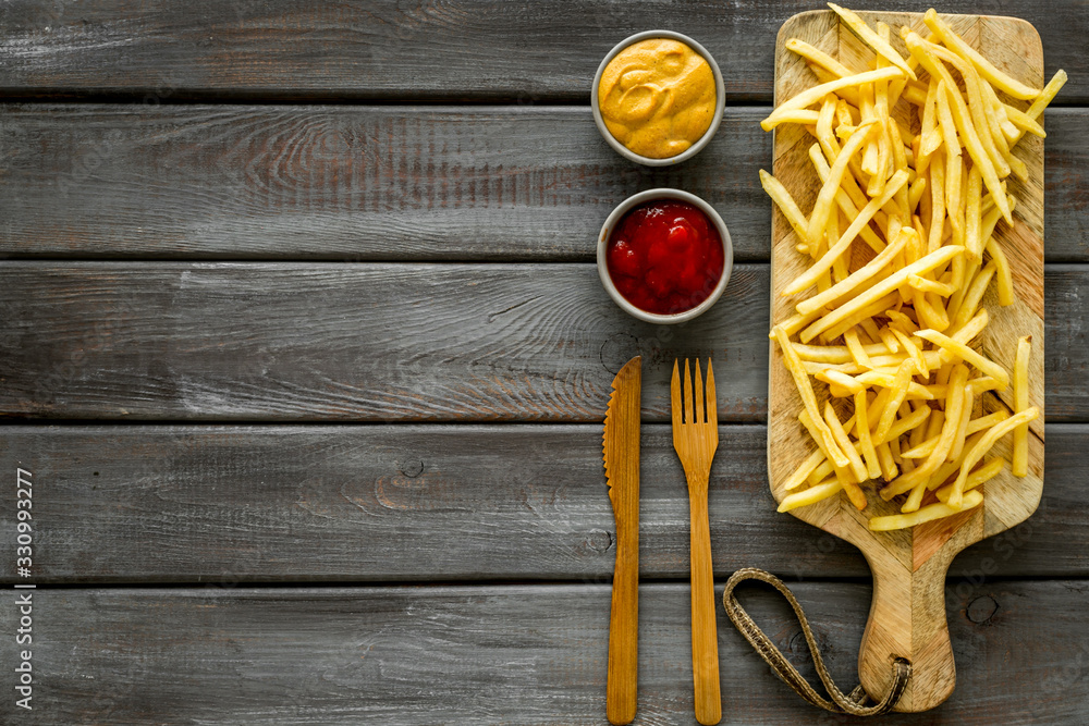 Fast food restaurant concept. French fries on cutting board near sauces on wooden table top-down cop