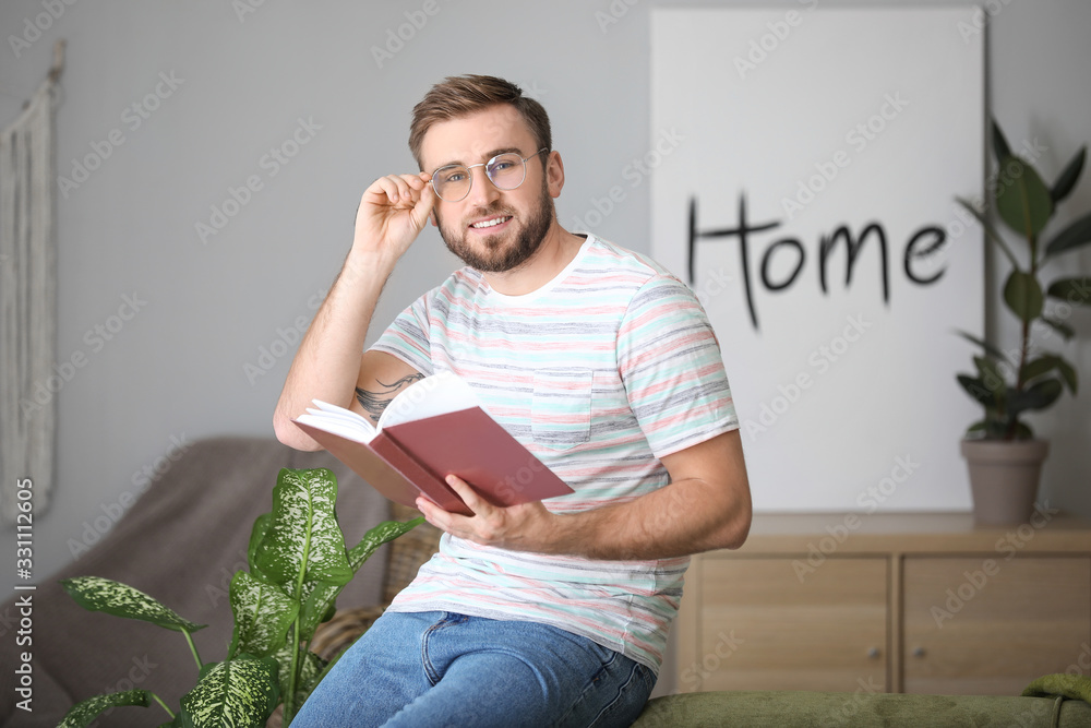 Young man with stylish eyeglasses reading book at home