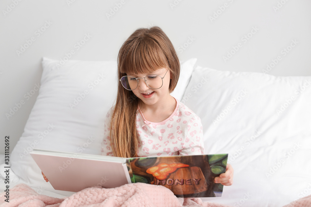 Cute little girl wearing glasses while reading book in bed