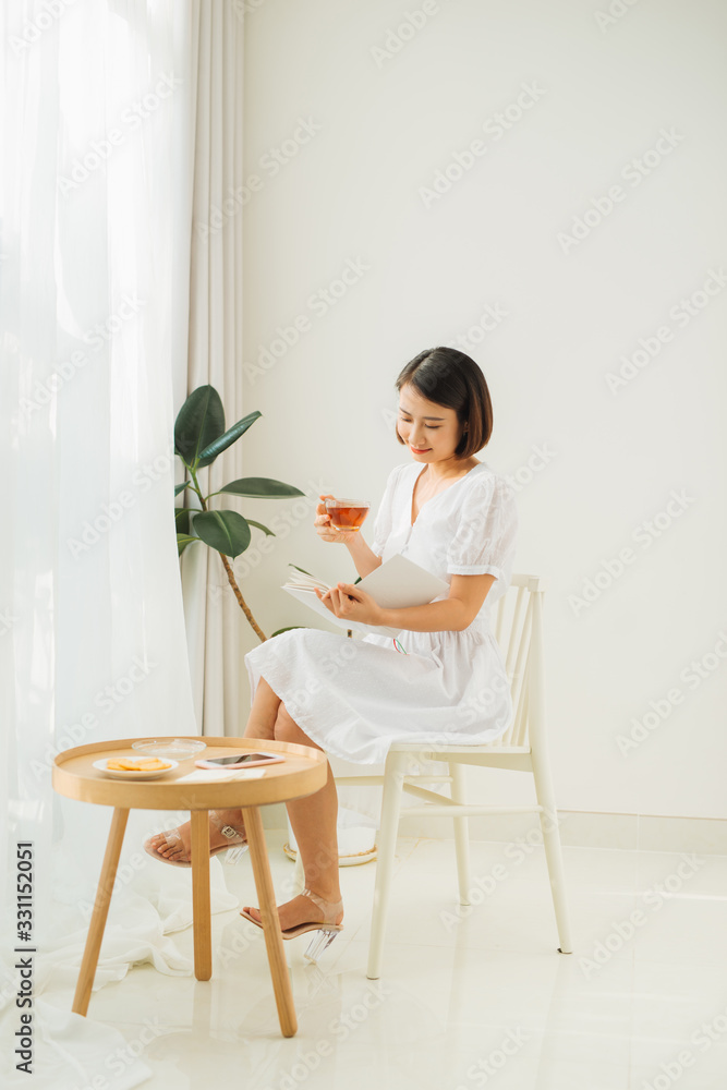 Young woman at home sitting near window relaxing in her living room reading book and drinking tea