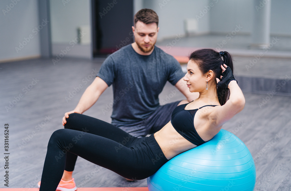 Trainer helping client workout on exercise ball at the gym. Sport and assistance concept. Doing exer