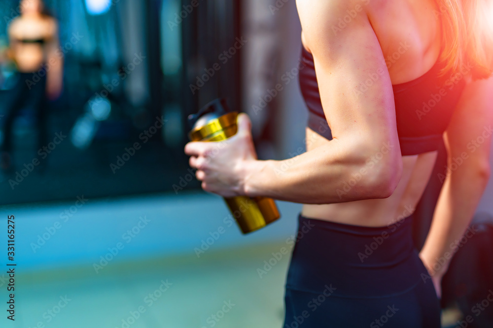 A girl with a bottle in the gym. Photo from the back. Cropped photo. Blurred background.