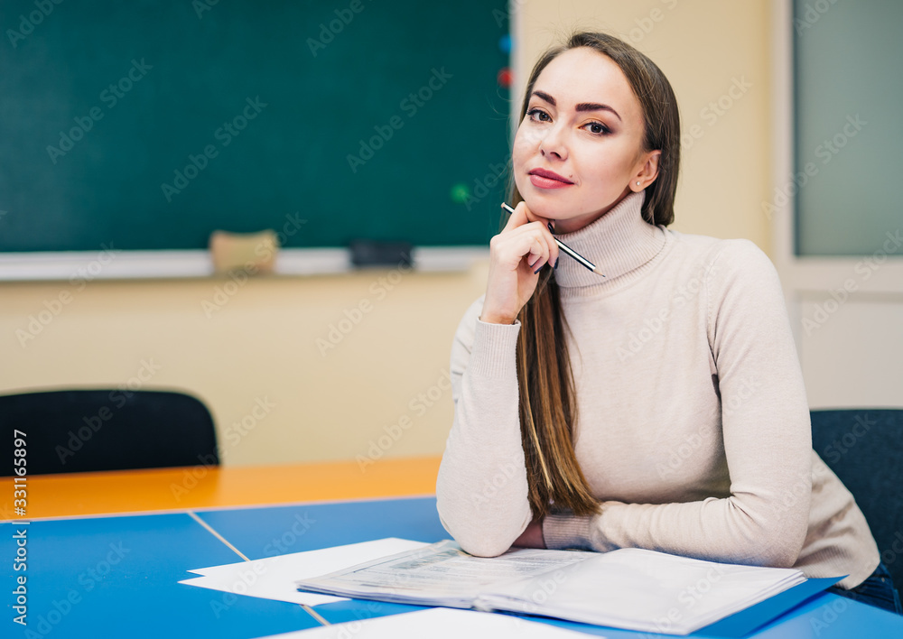 Beautiful young teacher with notes is sitting at table in classroom. Blackboard background