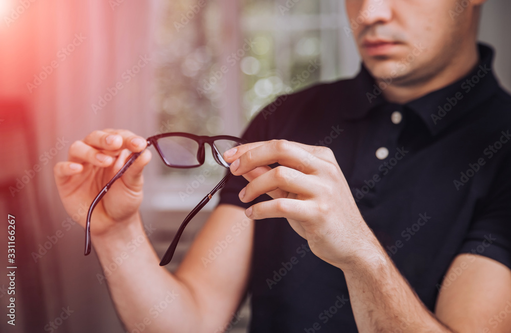 Young man holds stylish black-rimmed eyeglasses. Young male examines glasses in the room. Close-up