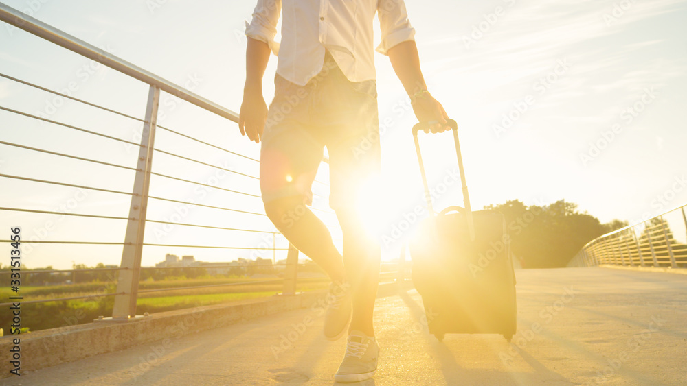 LOW ANGLE: Unrecognizable man walks across a bridge with his baggage at sunrise.