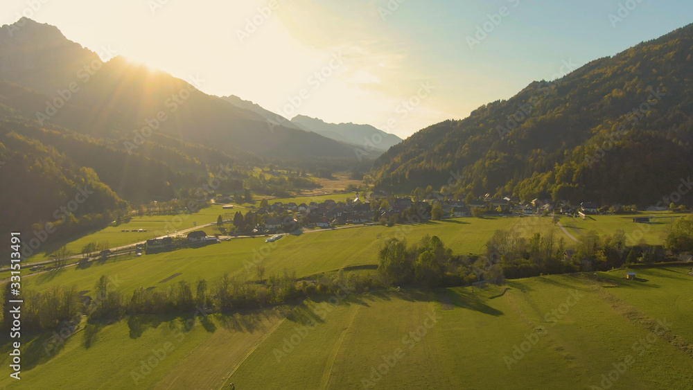 AERIAL: Golden summer sunbeams shine on a small village under a rocky mountain