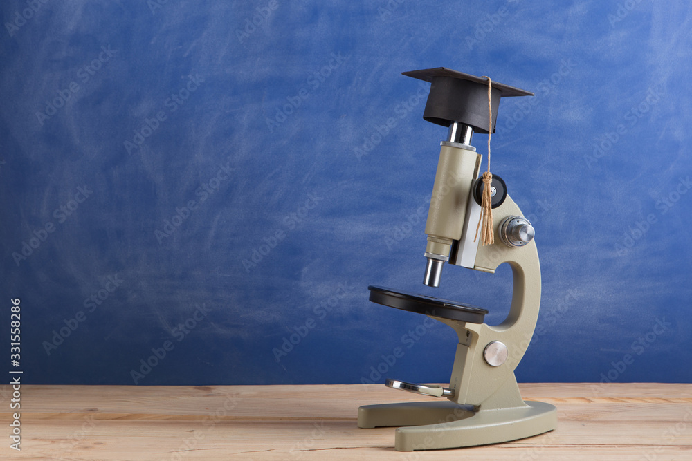 Education concept - microscope in a graduation hat on the desk in the auditorium. Blackboard backgro