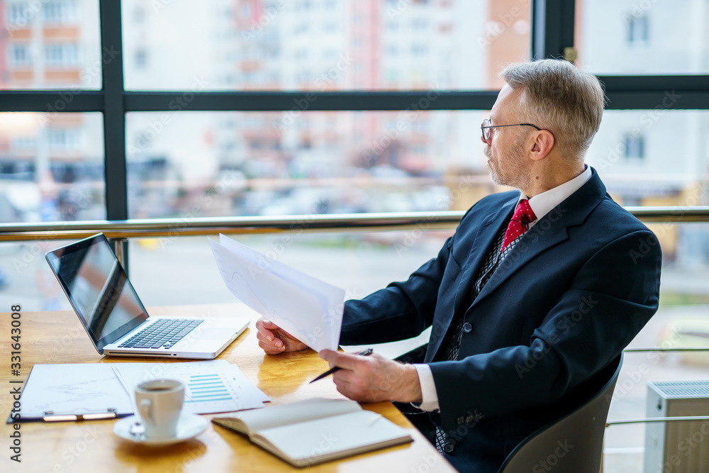 Thoughtful middle aged businessman in suit. Sitting with a laptop near the window. Man working with 