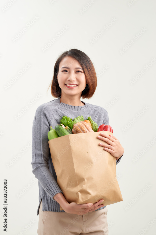 漂亮的年轻女人穿着围裙，手里拿着装满新鲜蔬菜的纸质购物袋，面带微笑