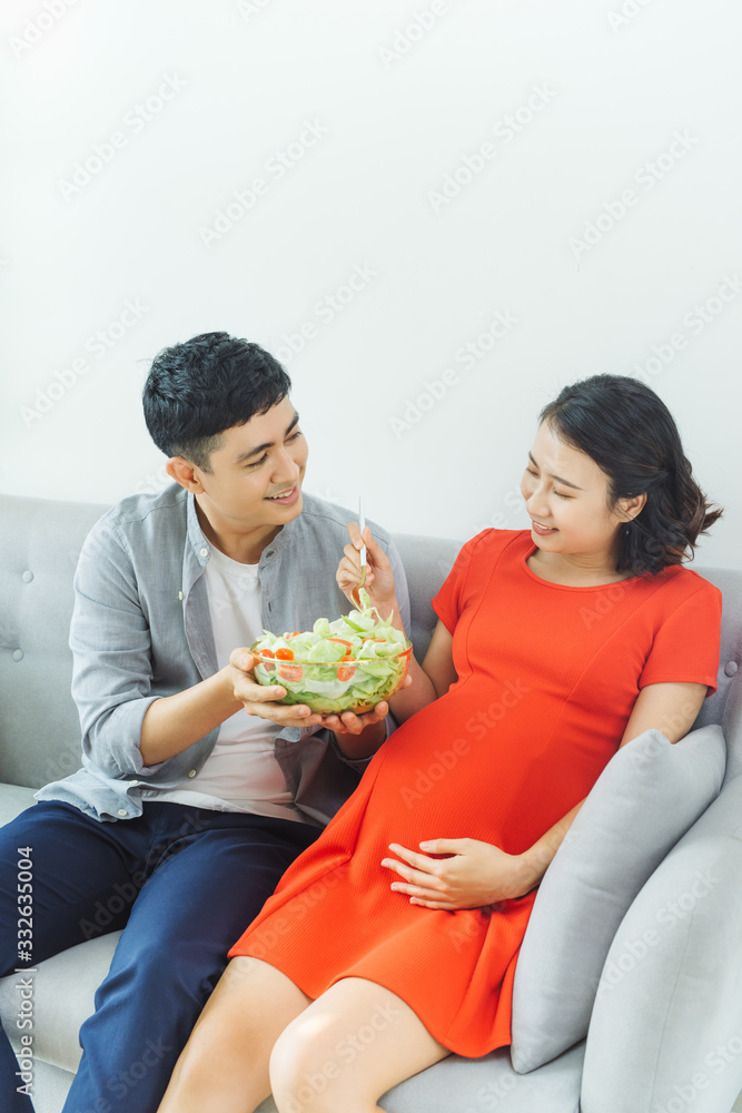 Lovely young pregnant couple relaxing on a couch together, woman having healthy breakfast