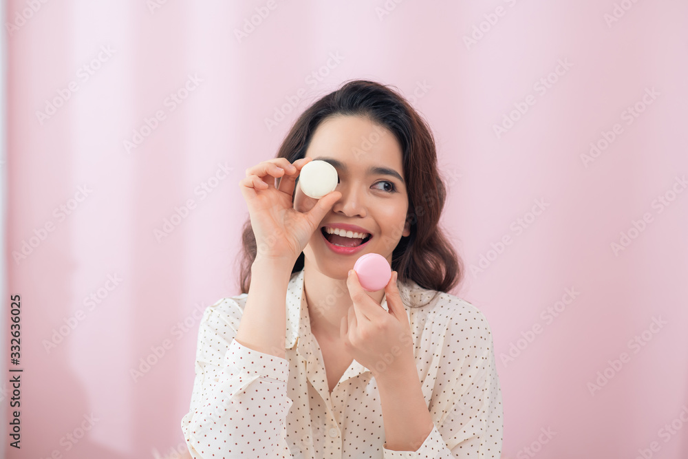 一位年轻的亚洲女性，在孤立的粉红色背景下，拿着五颜六色的马卡龙吃着它