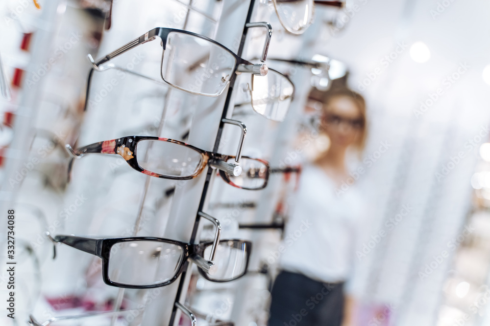 Optical shop. Optician suggest glasses. Woman standing with many eyeglasses in background.