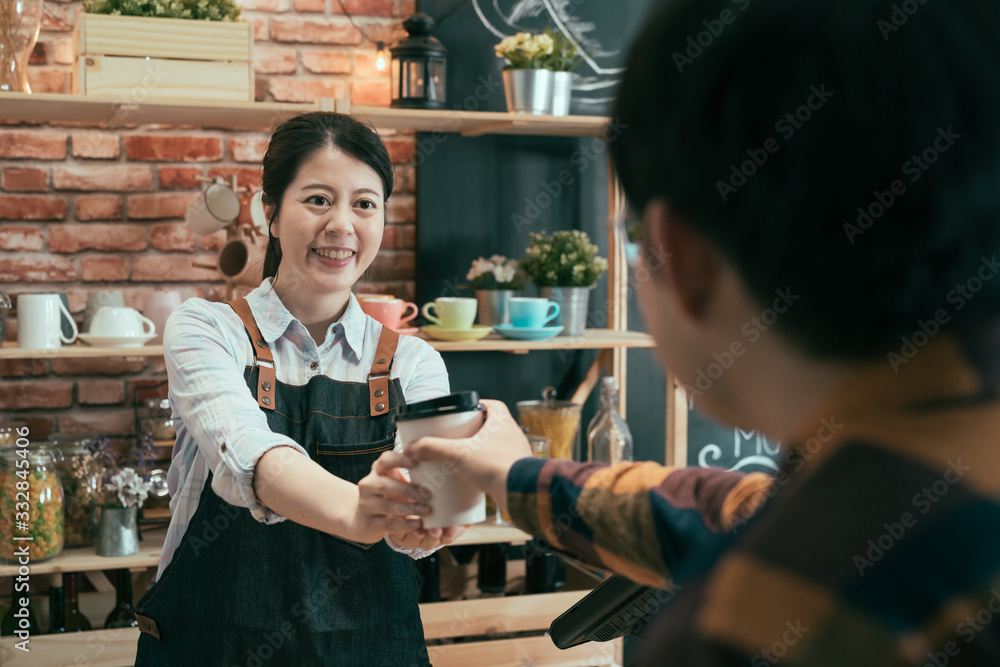 asian korean female barista in denim apron giving hot coffee in white takeaway paper cup to male cus