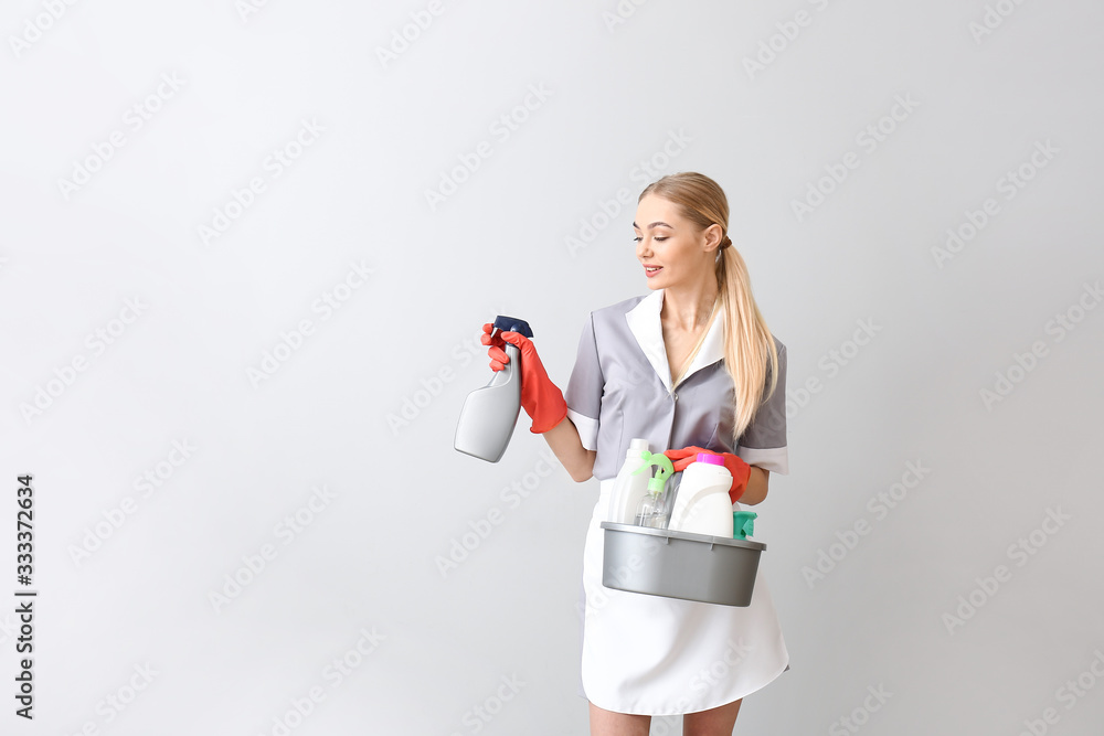 Beautiful young chambermaid with cleaning supplies on light background