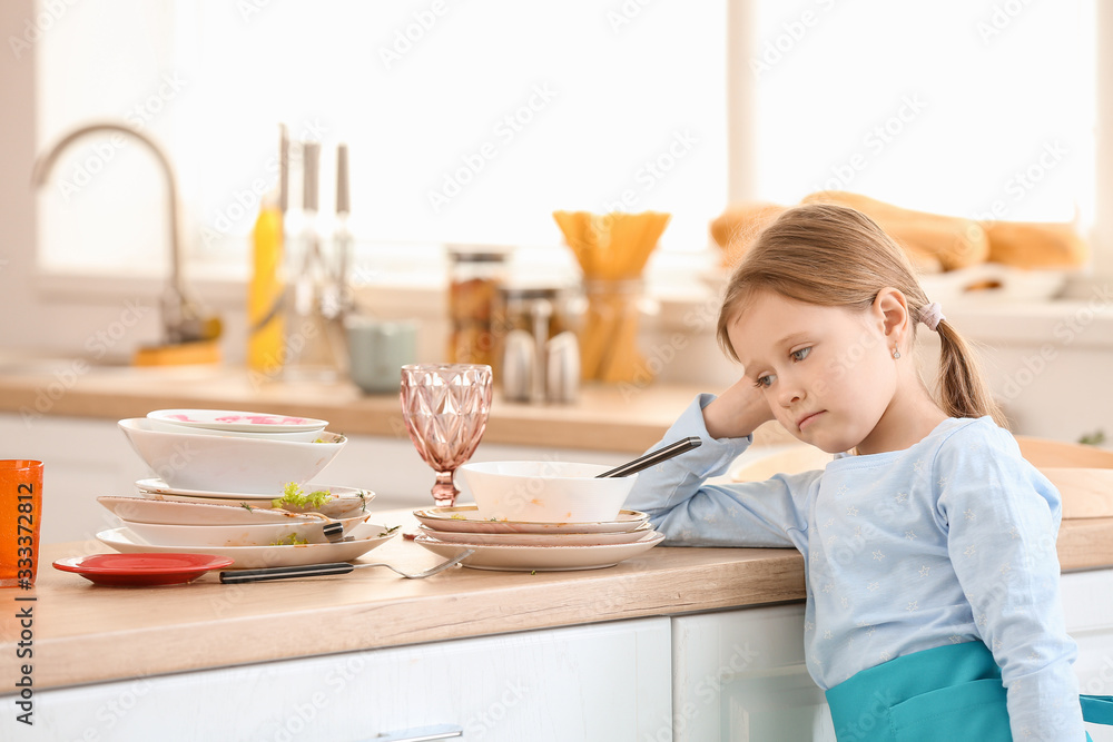 Reluctant little girl looking at dirty dishes in kitchen