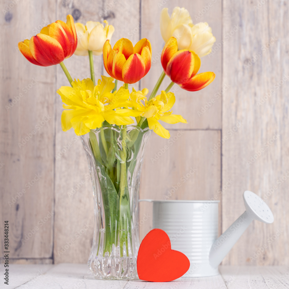 Tulip flower arrangement in glass vase with heart greeting, watering can decor on wooden table backg