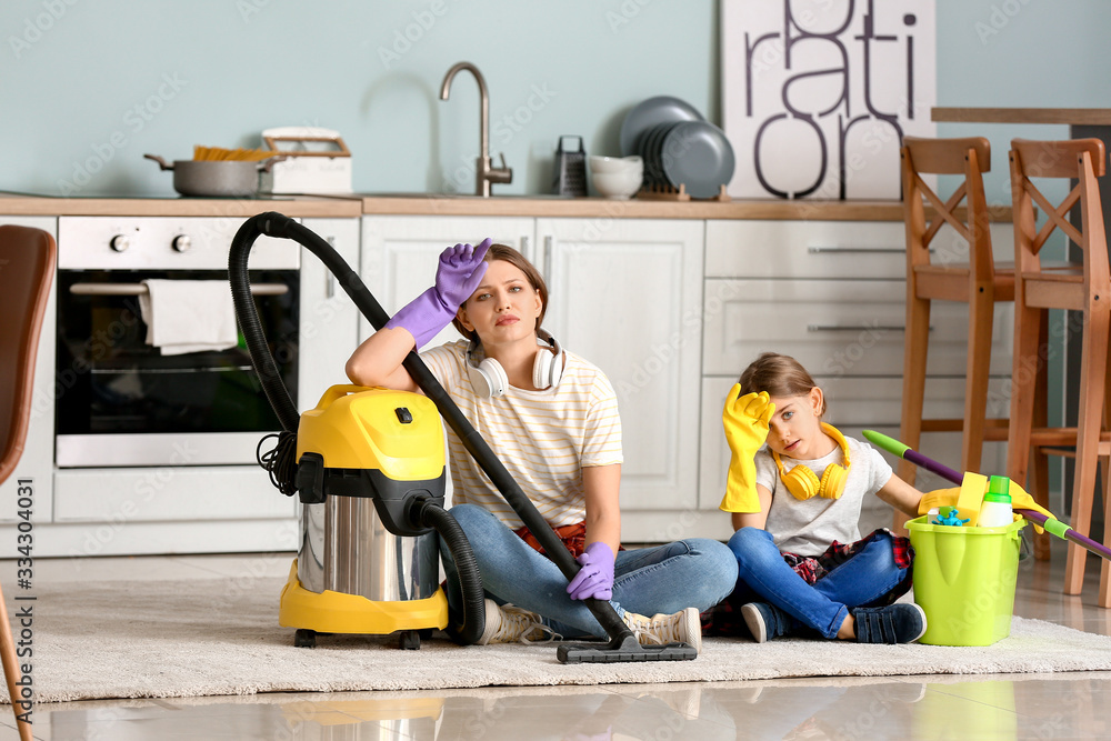 Tired woman and her little daughter with vacuum cleaner in kitchen